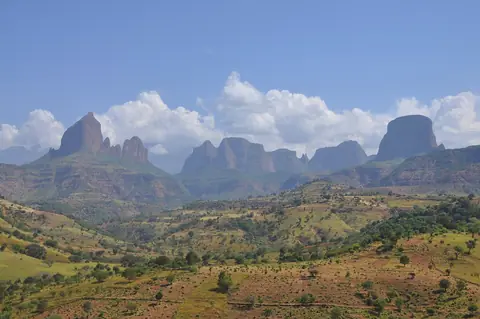 Simien-bjergenes stejle klippetinder bag grønne, dyrkede bakker under blå himmel med hvide skyer