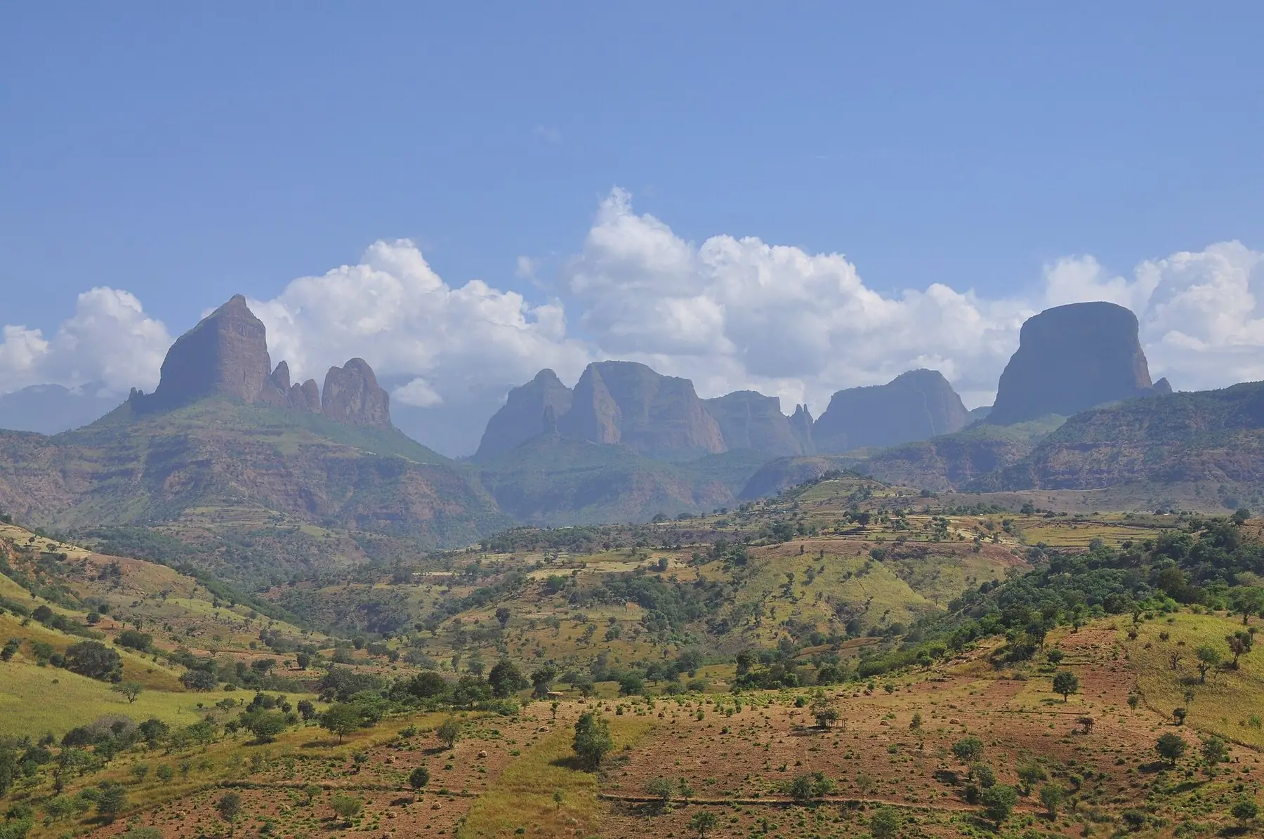 Simien-bjergenes stejle klippetinder bag grønne, dyrkede bakker under blå himmel med hvide skyer