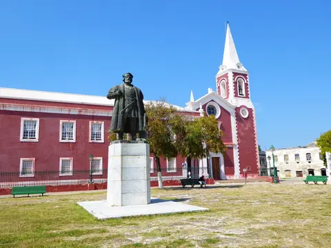 Rødmalet palads og en kirke med spidst tårn bag en statue på en græsplads på Mozambique-øen