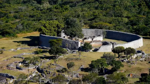 Den høje, afrundede stenmur om Great Enclosure i ruinbyen Great Zimbabwe mellem træer og græs