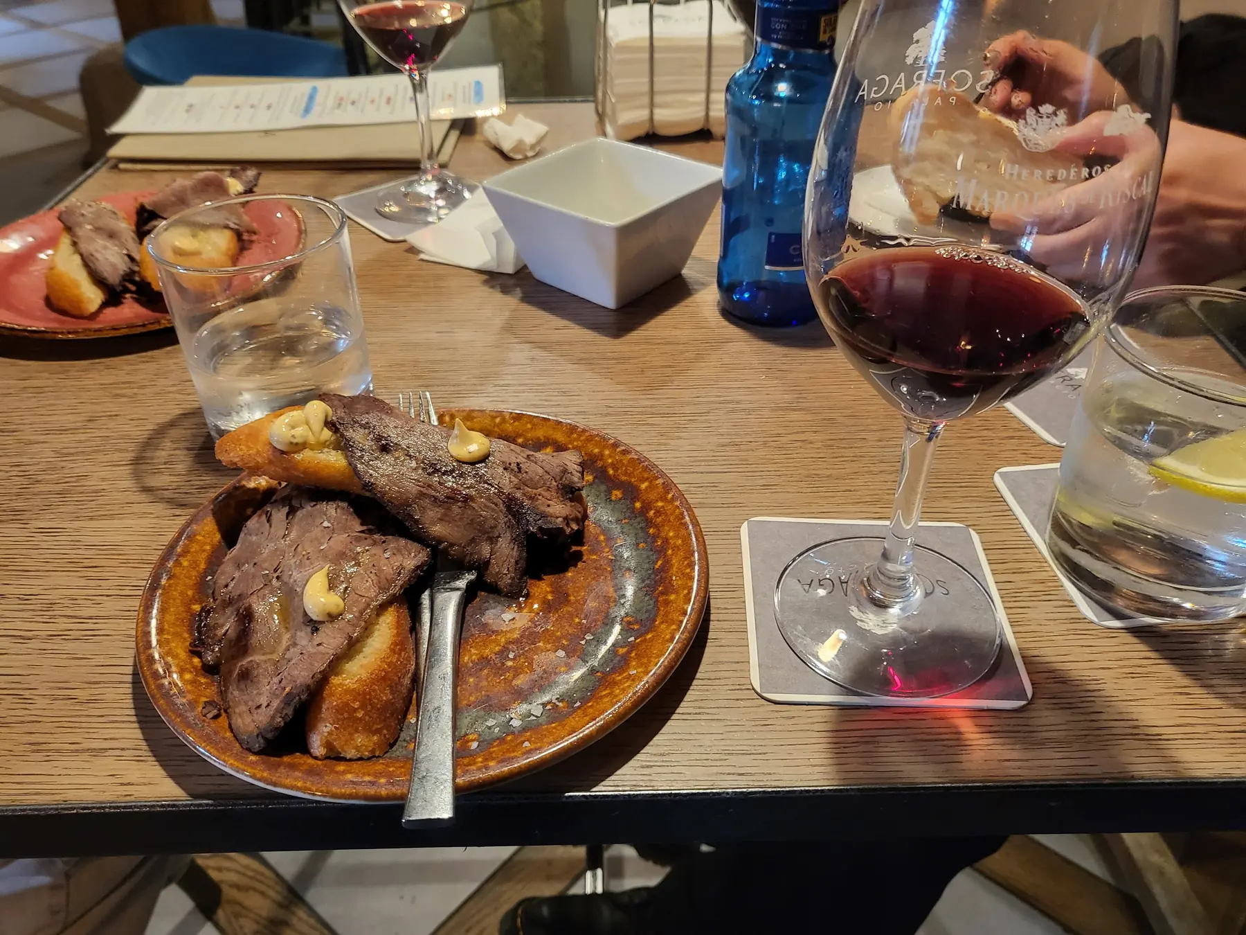 Et glas rødvin ved siden af en tapa med oksekød på brød på et træbord i en spansk bar