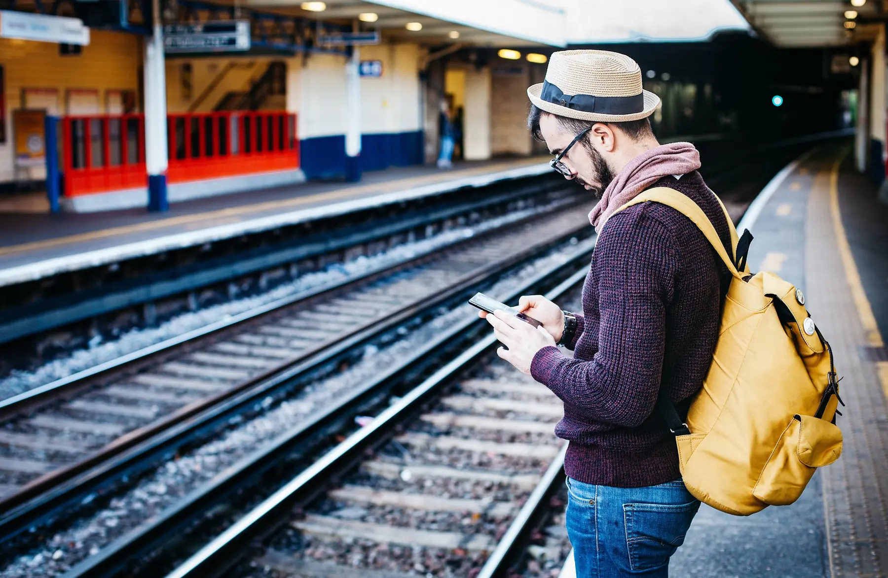 En rejsende med gul rygsæk og stråhat står på en perron og kigger på sin telefon