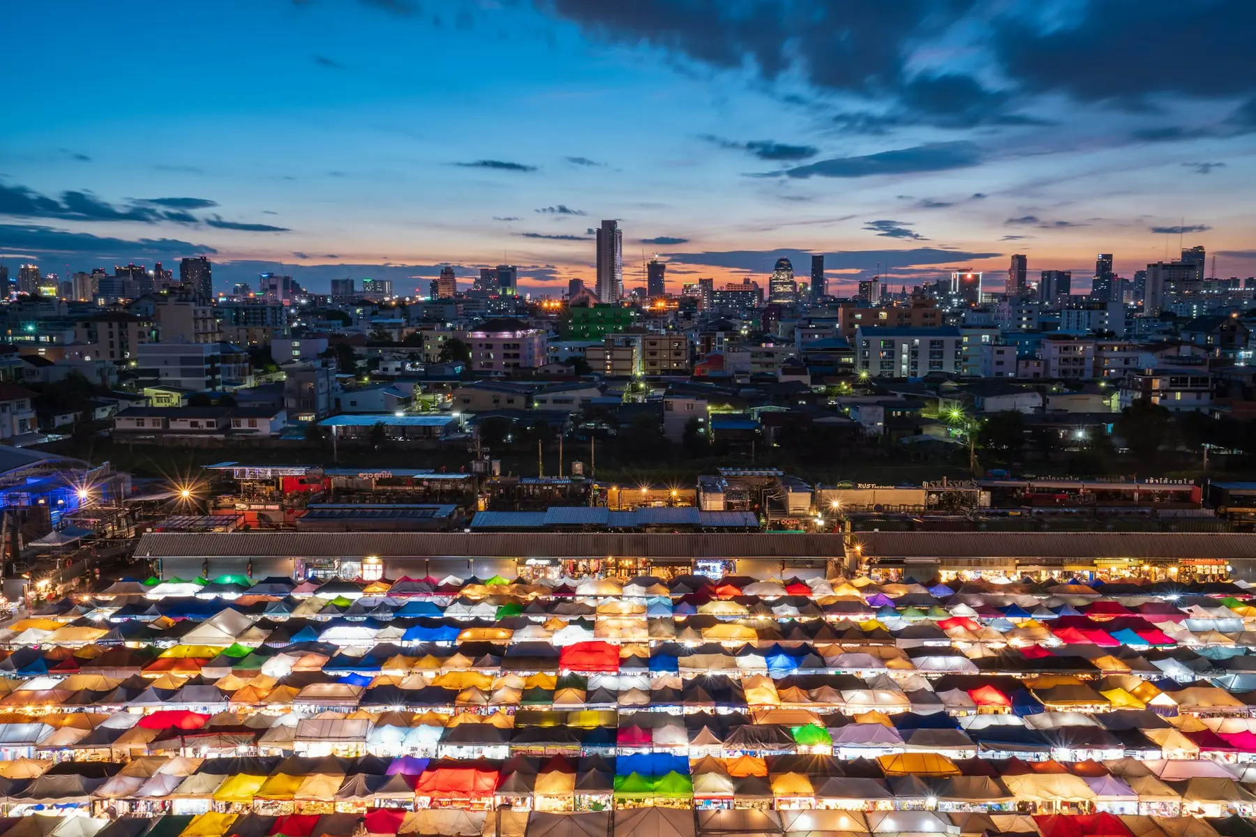 Hundredvis af farverige boder på et nattemarked set oppefra med Bangkoks skyline i skumringen