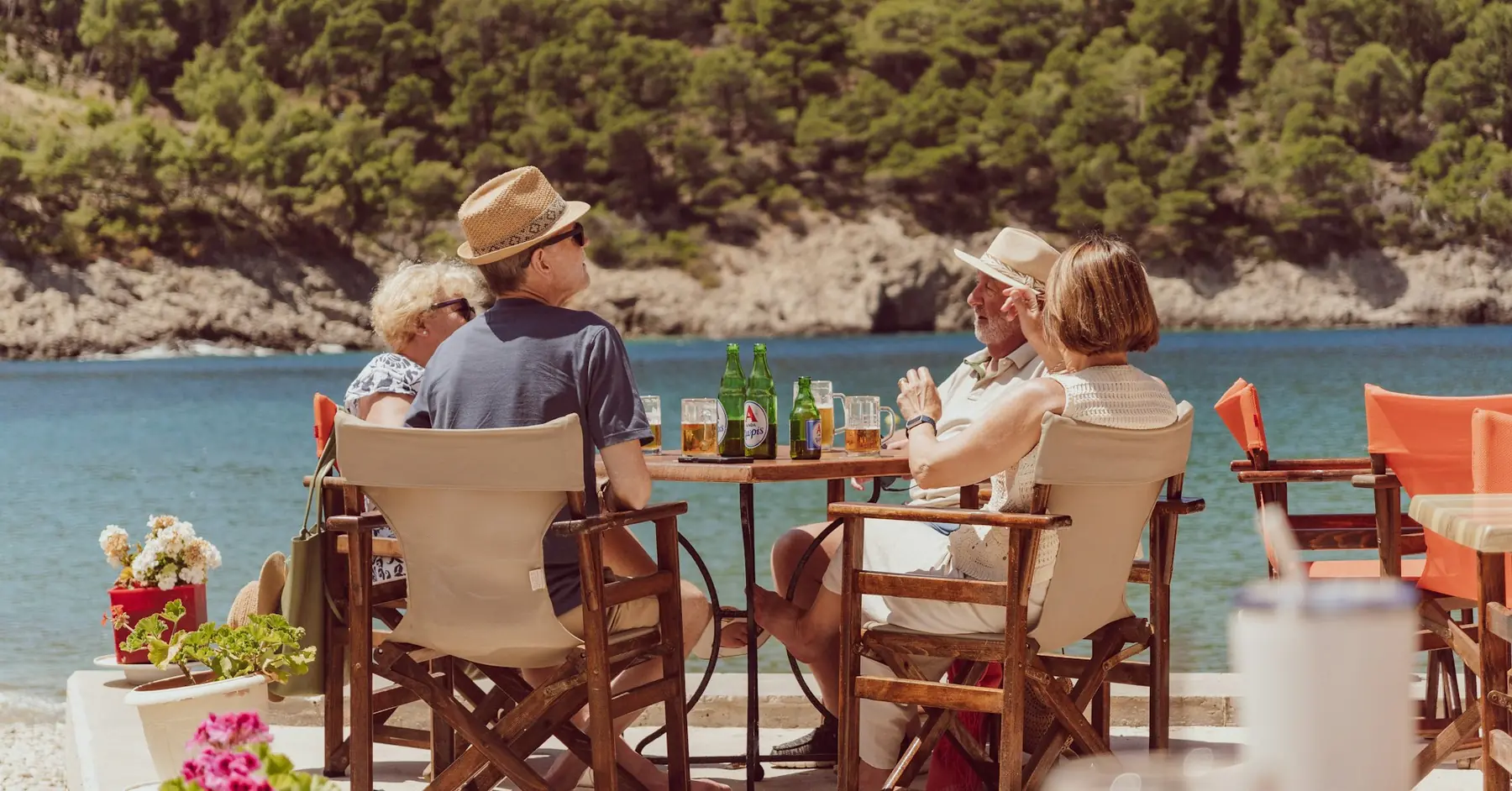 To ældre par sidder ved et bord med øl på en taverna ved en klippebugt med klart vand