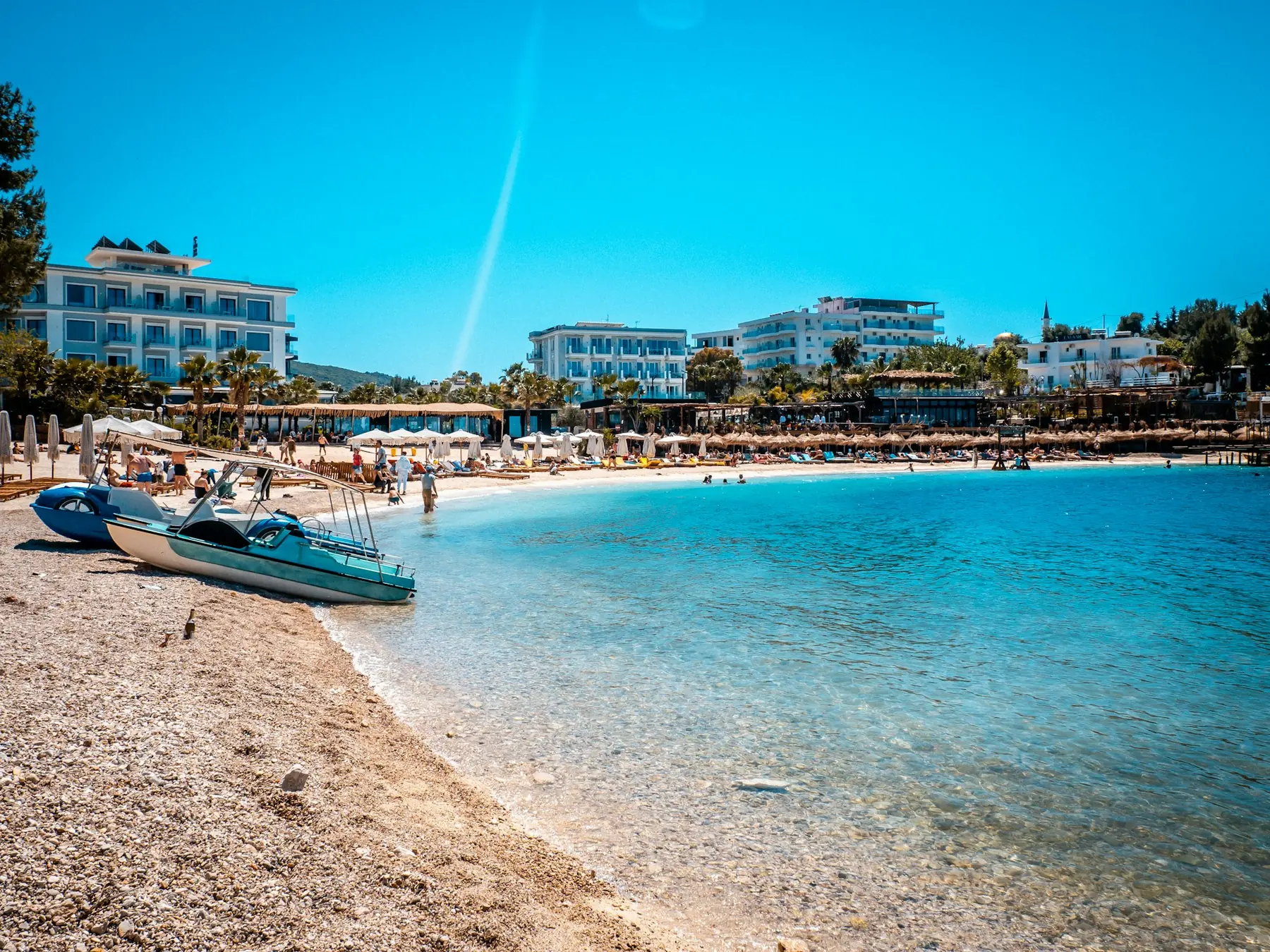 Klart turkist vand ved en rullestensstrand med parasoller, pedalbåde og hvide hoteller i Albanien