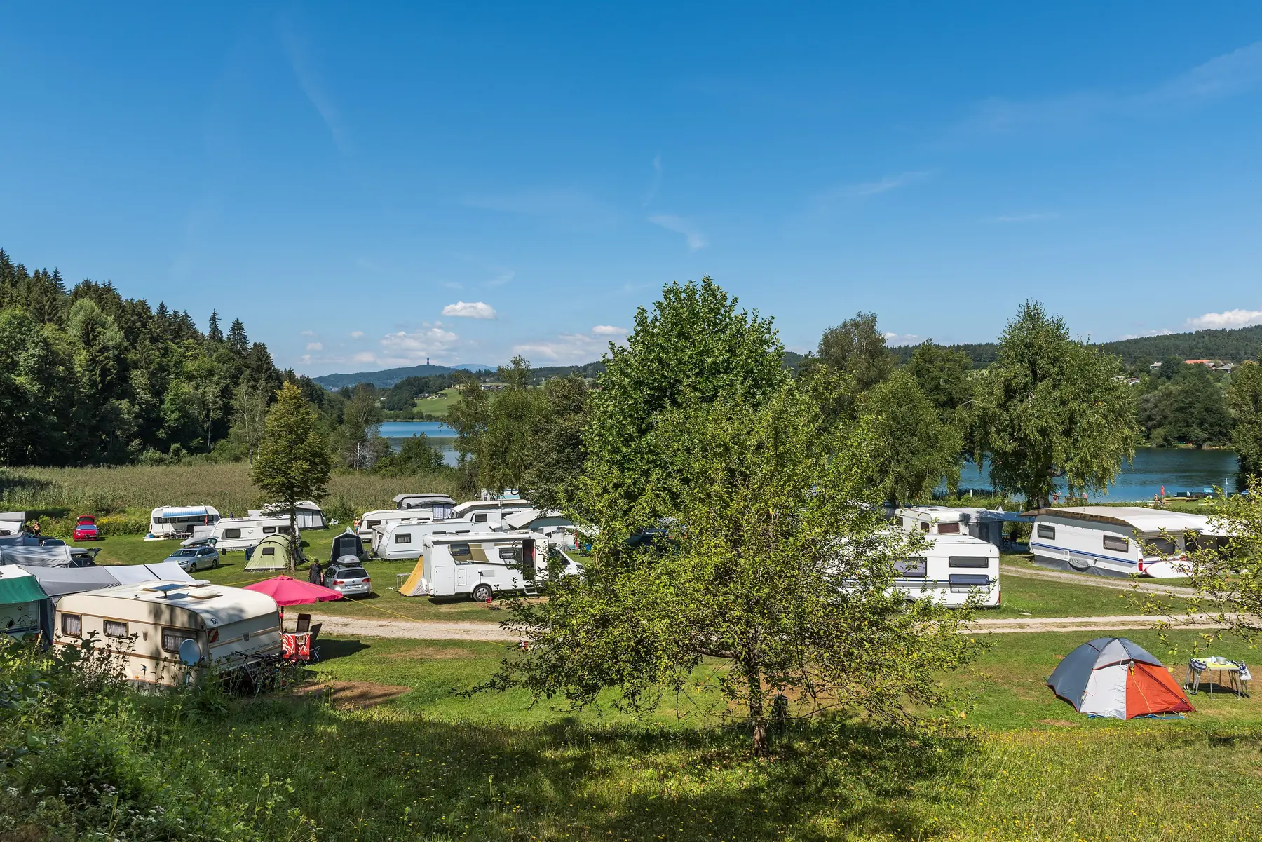 Campingvogne, autocampere og telte på en grøn campingplads ved en sø omgivet af skov i Kärnten