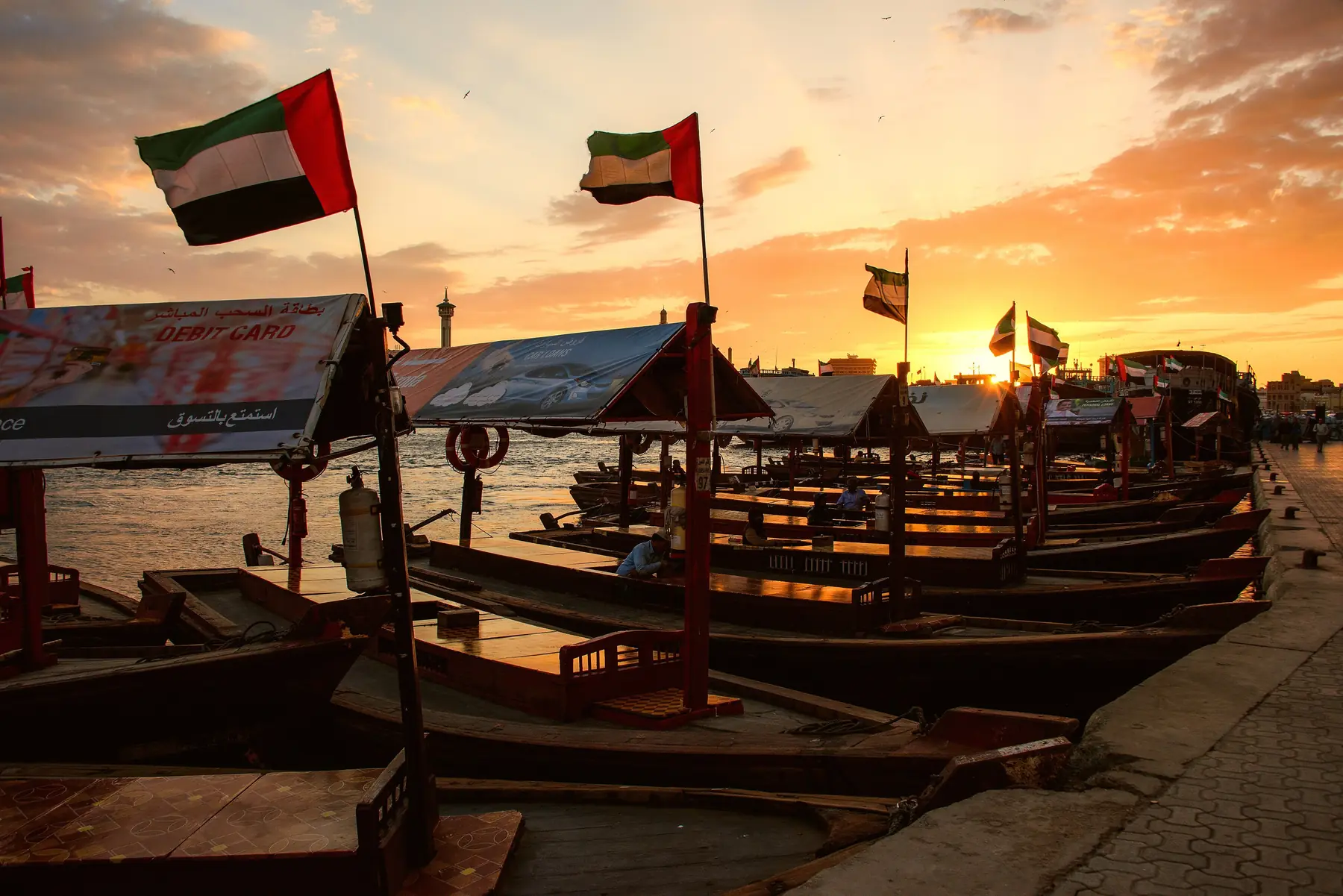 Fortøjede træfærger af abra-typen med flag ved Dubai Creek i solnedgang under en orange himmel