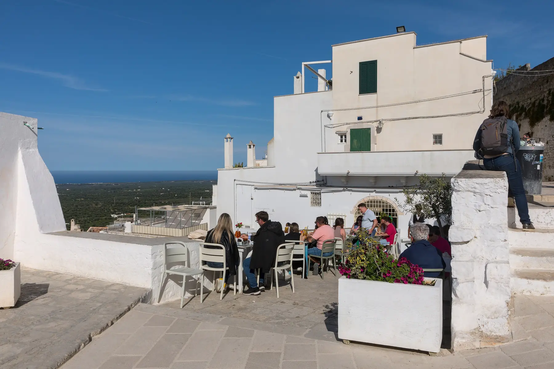 Gæster ved borde på en caféterrasse mellem hvidkalkede huse i Ostunis gamle bydel med udsigt over sletten mod havet