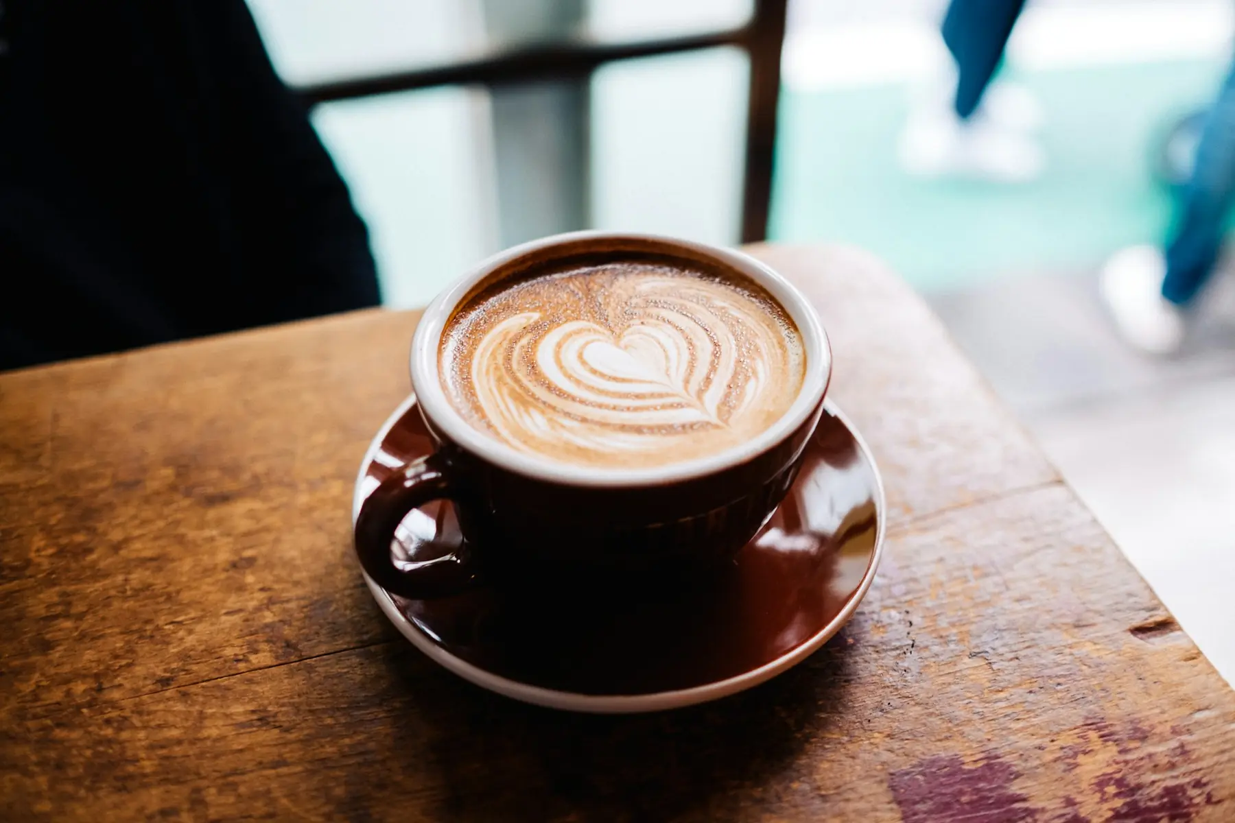 En cappuccino med latte art i en brun kop på et slidt træbord ved vinduet på en café