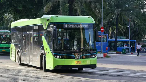 En grøn elbus kører gennem et kryds med palmer og blå bybusser i baggrunden i Vietnam