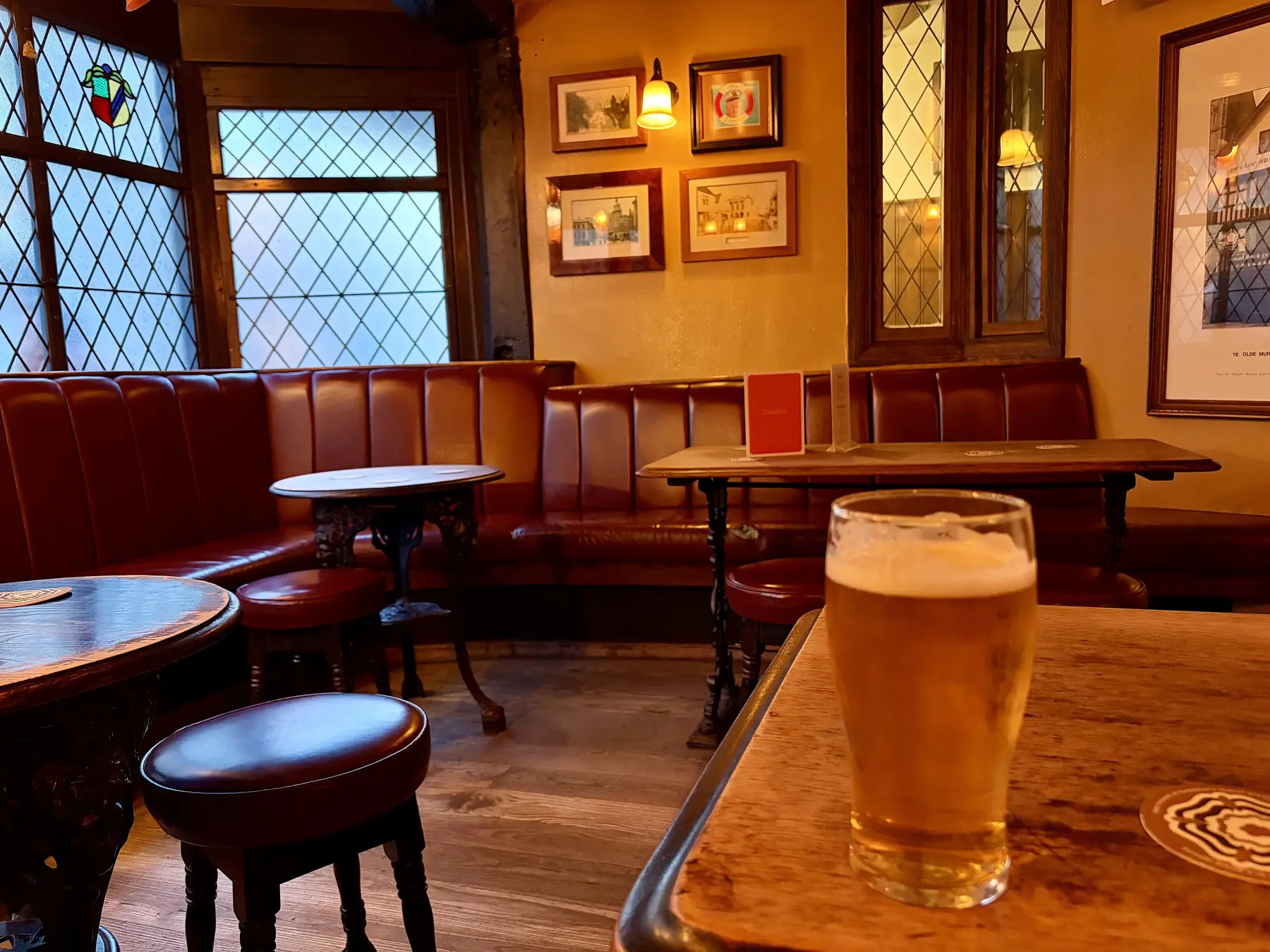 Et glas lys fadøl på et træbord i en gammel pub med læderbænke, blyindfattede ruder og billeder på væggen