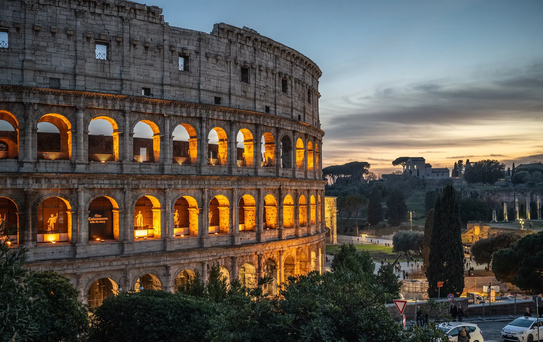 Colosseum i Rom med oplyste buegange i skumringen og pinjer under en blå og gylden himmel