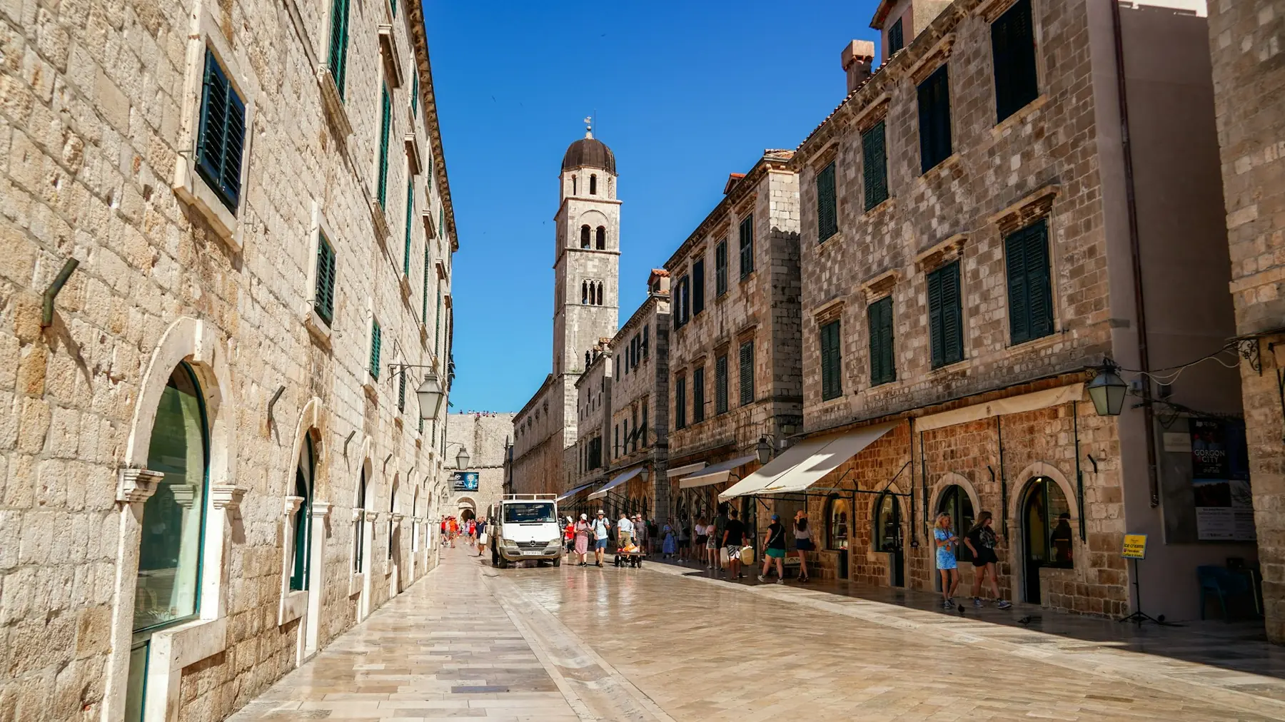 Turister på den blanke brolagte hovedgade Stradun i Dubrovniks gamle bydel med franciskanerklosterets klokketårn