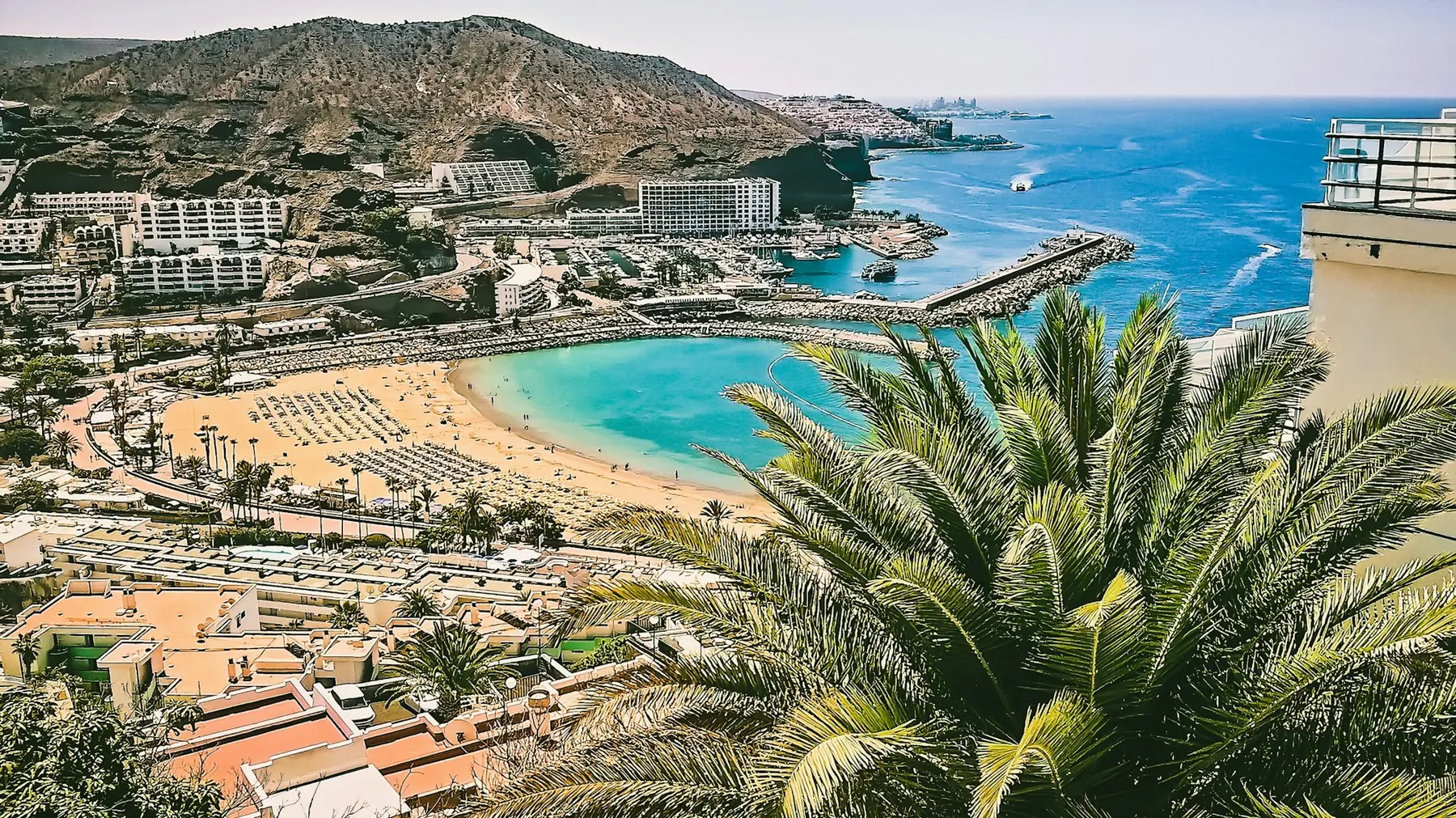 Halvmåneformet sandstrand med turkisblåt vand, hoteller på de tørre bjergskråninger og palmeblade i forgrunden