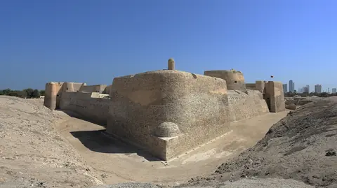 Den gamle sandstensfæstning Qal'at al-Bahrain under klar blå himmel med højhuse i det fjerne