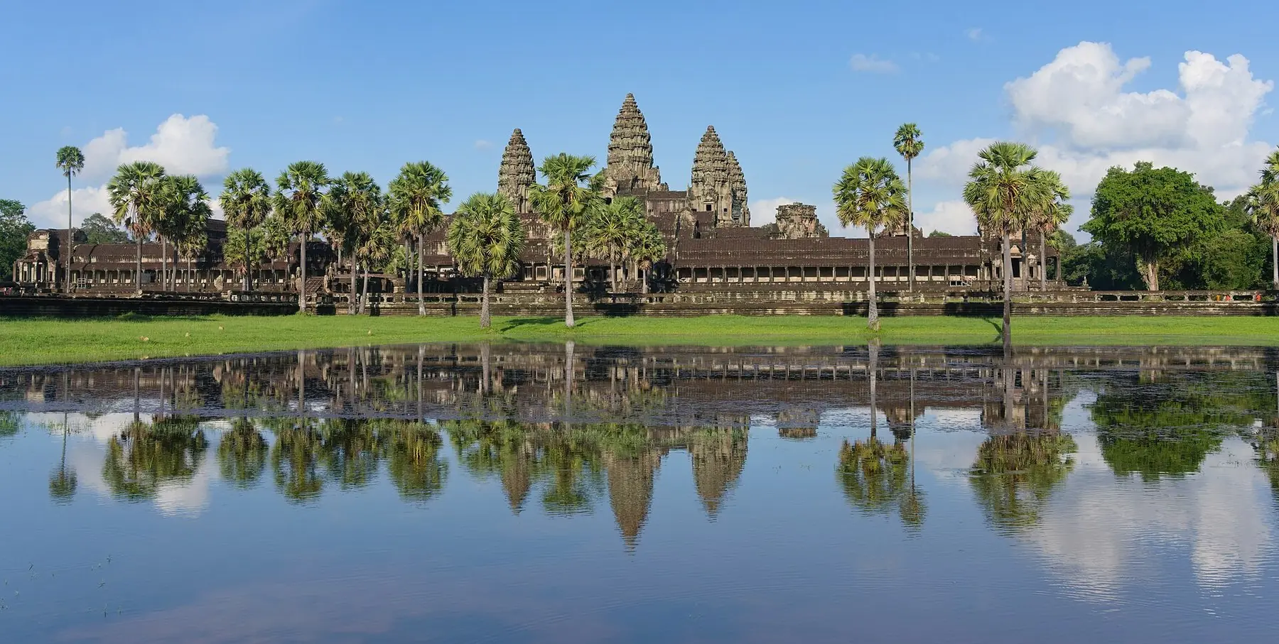 Angkor Wats tempeltårne og palmer, der spejler sig i en stille dam under blå himmel