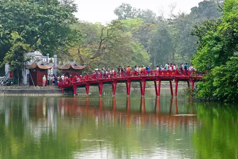Den røde Huc-bro over Hoan Kiem-søen i Hanoi med besøgende og grønne træer der spejler sig i vandet