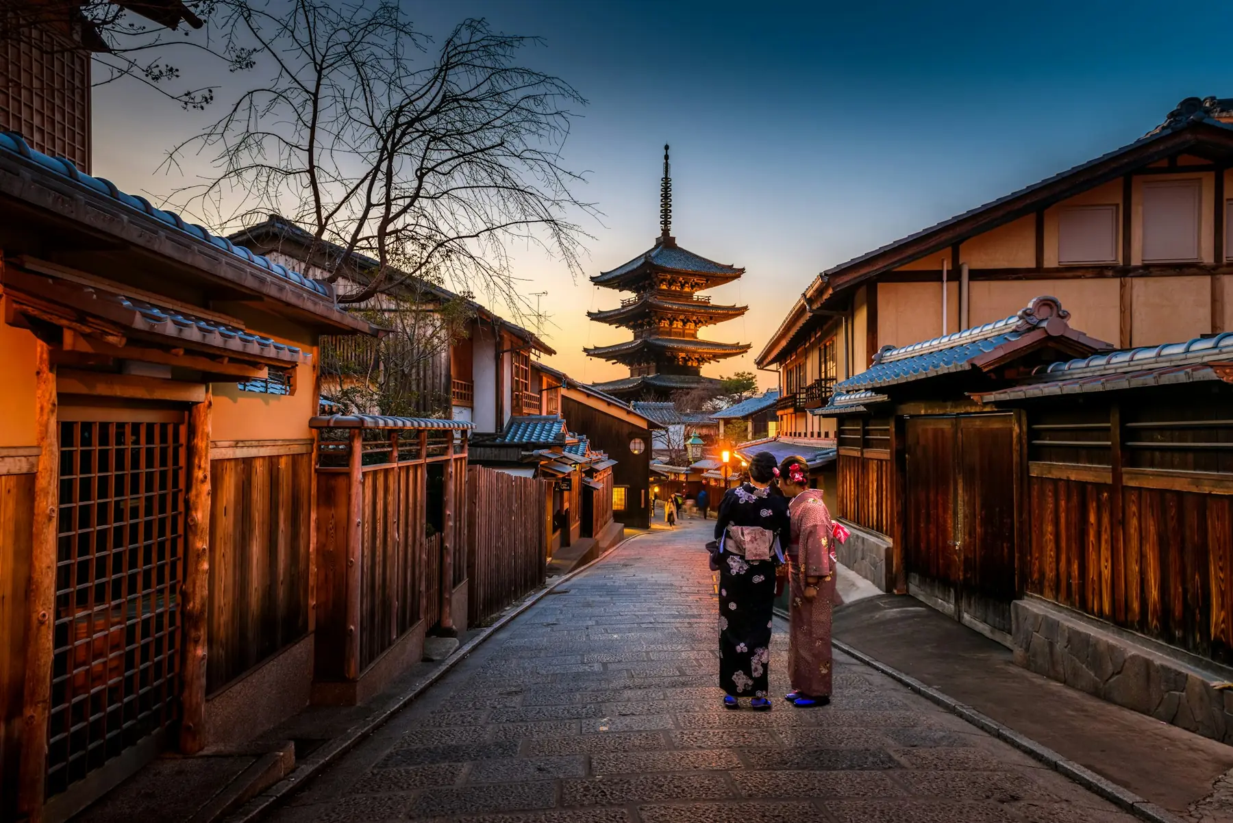 To kvinder i kimono på en brolagt gade med gamle træhuse og Yasaka-pagoden i Kyoto ved solnedgang