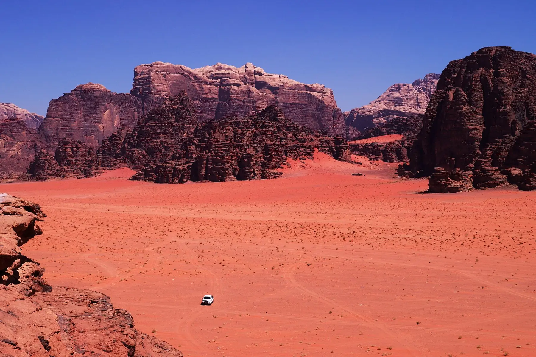 Rød sandørken mellem høje sandstensbjerge i Wadi Rum, set fra oven med en enlig terrængbil