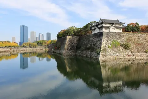 Borgmuren og et tårn ved Osaka-borgens voldgrav med højhuse og efterårsløv der spejler sig i vandet