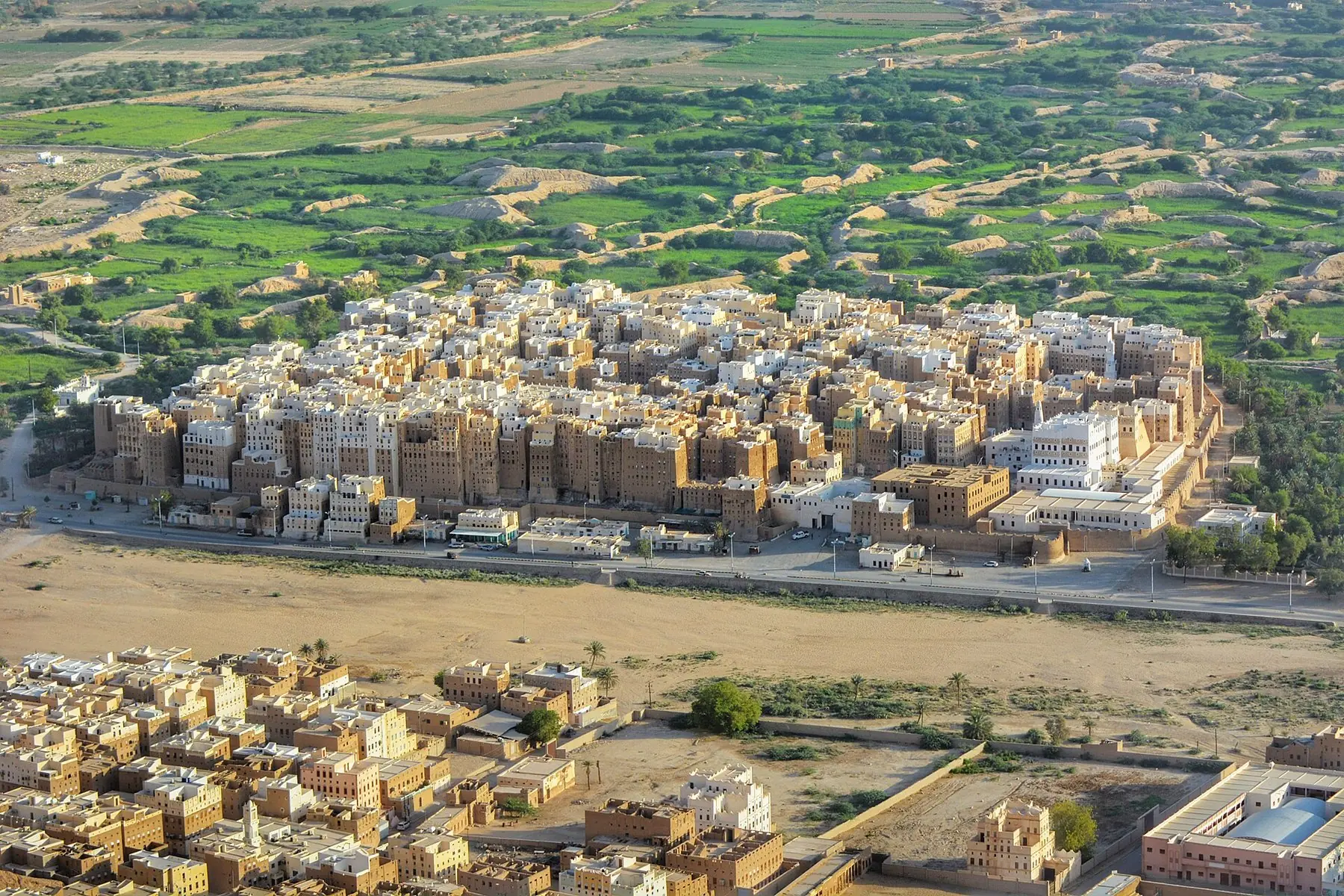 Den muromkransede by Shibam med tætpakkede lerhøjhuse omgivet af grønne marker i Hadramaut-dalen, set fra oven