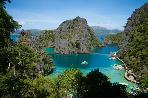 Kalkstensklipper og turkis vand med udriggerbåde ved en træbro set fra udsigtspunktet ved Kayangan Lake på Coron