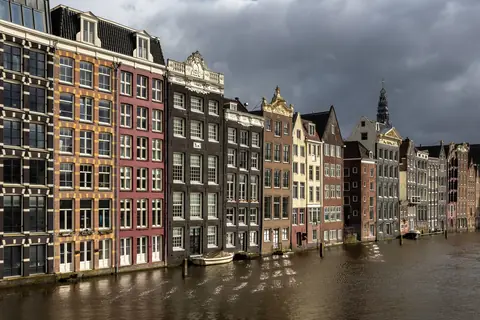 Smalle, farverige kanalhuse ved Damrak i Amsterdam i aftensol under mørke skyer