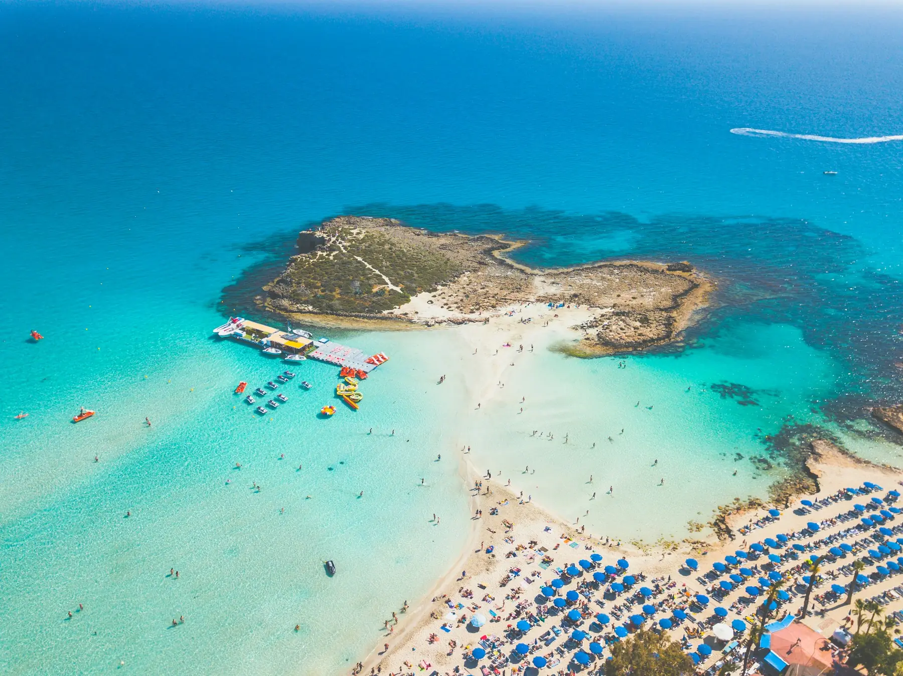 Luftfoto af Nissi-stranden ved Ayia Napa på Cypern med lavt turkist vand, en lille klippeø og parasoller