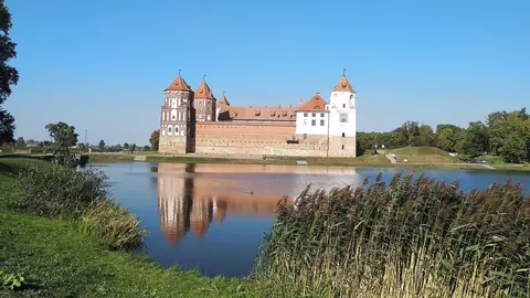 Mir-slottet med røde tage og tårne spejlet i en sø omkranset af siv i Hviderusland