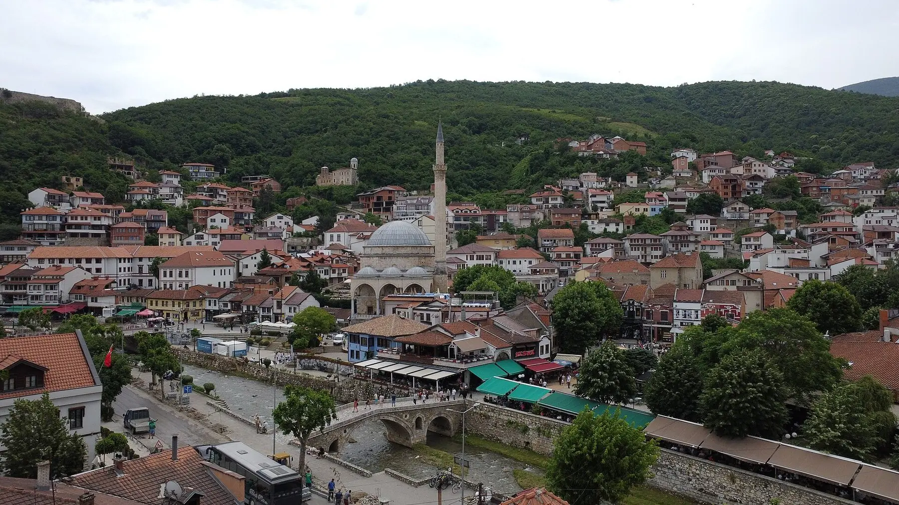 Moské med kuppel og minaret ved floden og en stenbro, omgivet af røde tage og skovklædte bjerge i Prizren