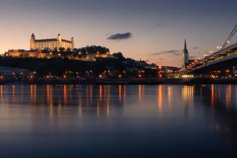 Bratislava Slot og Sankt Martins katedral oplyst i skumringen og spejlet i Donau ved broen SNP