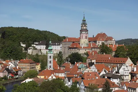 Røde tegltage og slotstårnet i Český Krumlov mellem grønne bakker under blå himmel