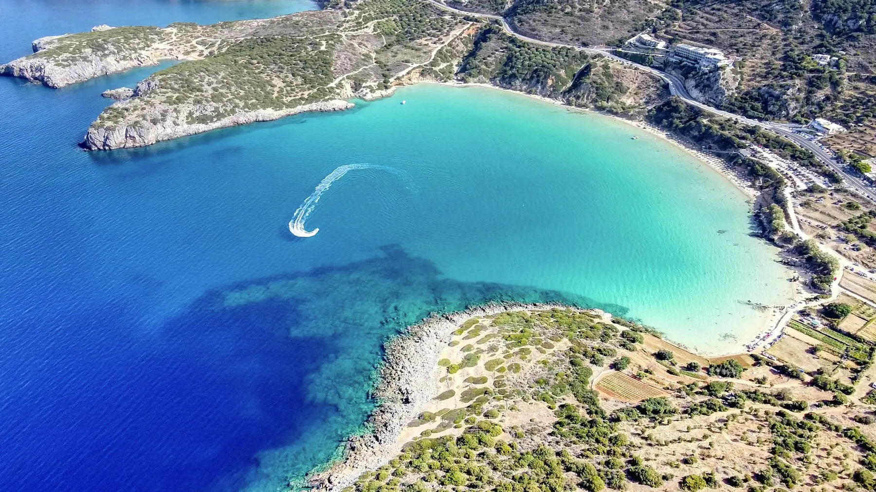 Luftfoto af en turkis badebugt med sandstrand mellem klippenæs og grønne skråninger på Kretas kyst
