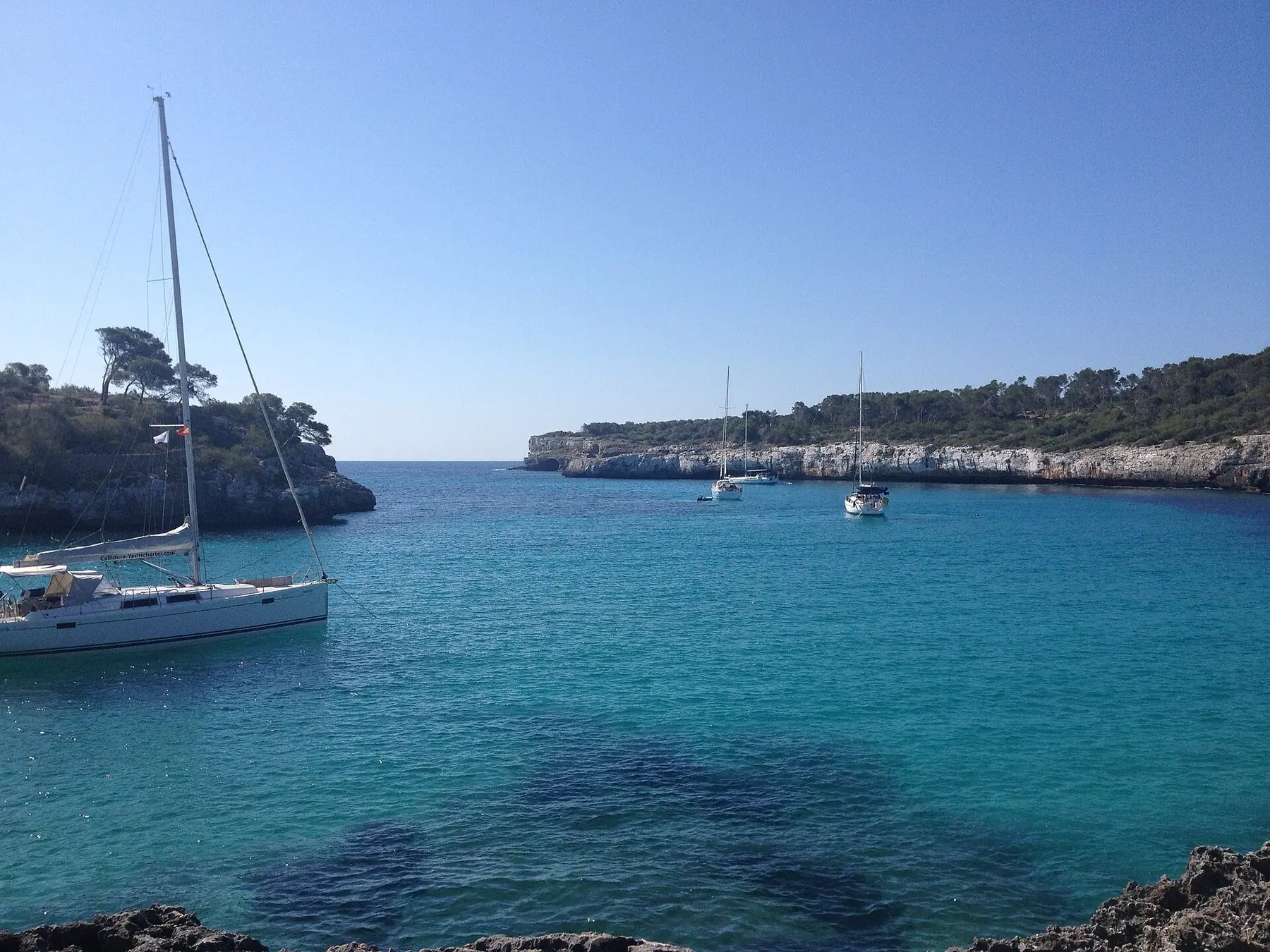 Sejlbåde i turkist vand mellem fyrreklædte klipper ved Cala Mondragó på Mallorca