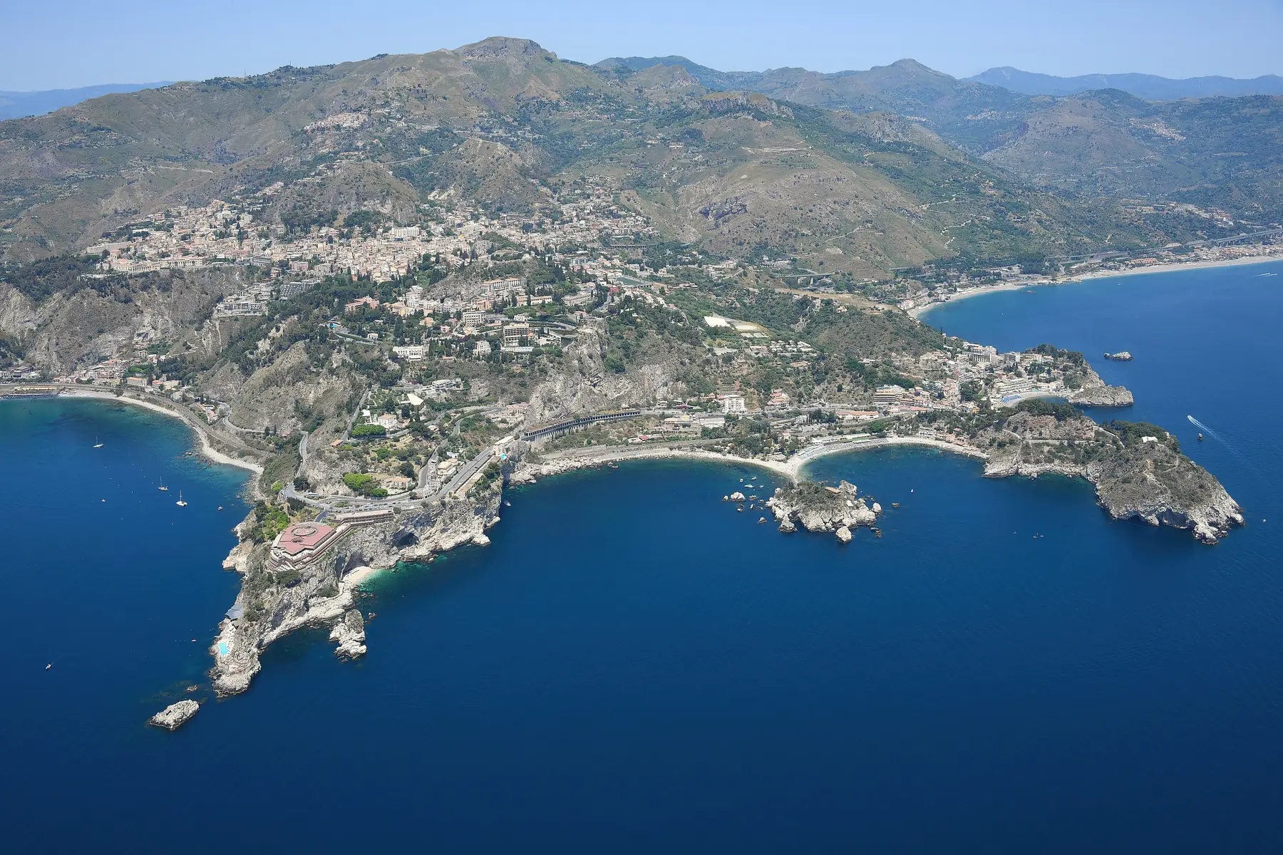 Luftfoto af kysten ved Taormina på Sicilien med små bugter, øen Isola Bella og bjergene bag