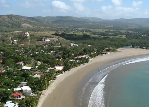 Den buede sandstrand i San Juan del Sur med huse mellem træerne og grønne bakker