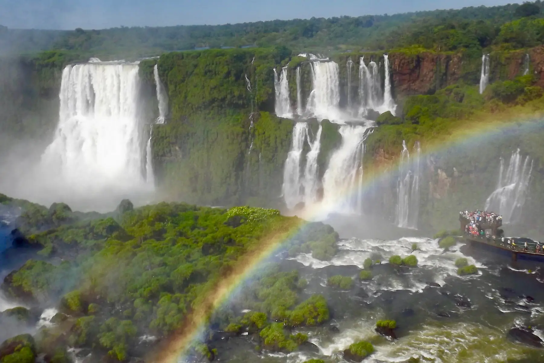 Iguaçu-vandfaldene styrter ned mellem grøn regnskov med en regnbue og en udsigtsbro fuld af besøgende